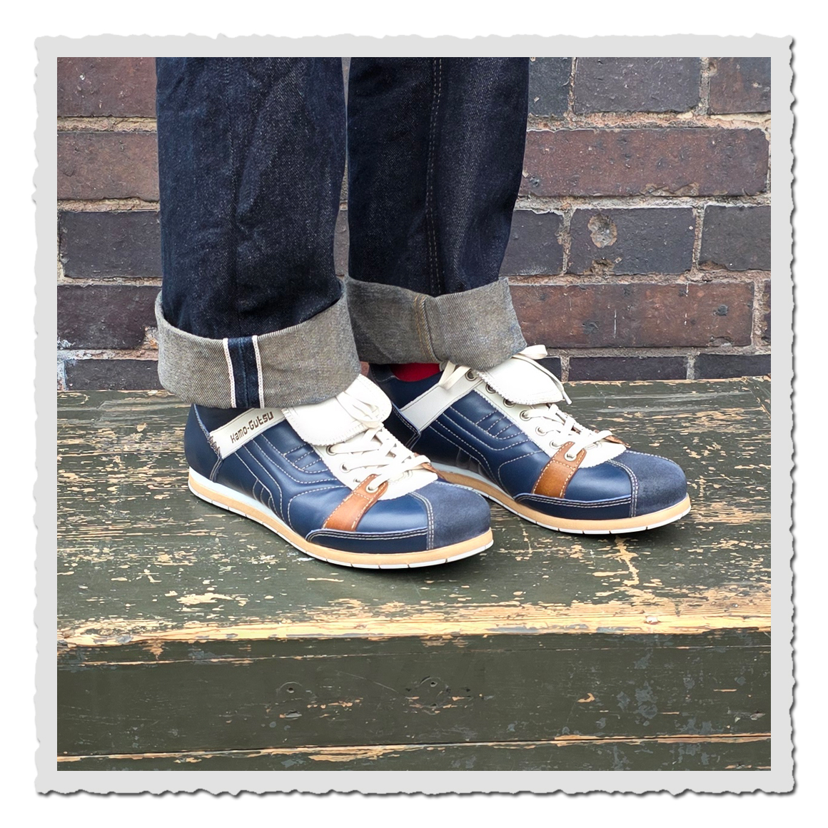 Dunkelblaue Schnürschuhe mit heller Lasche an einer Person mit dunklen Jeans vor Backsteinwand auf Holzpodest.